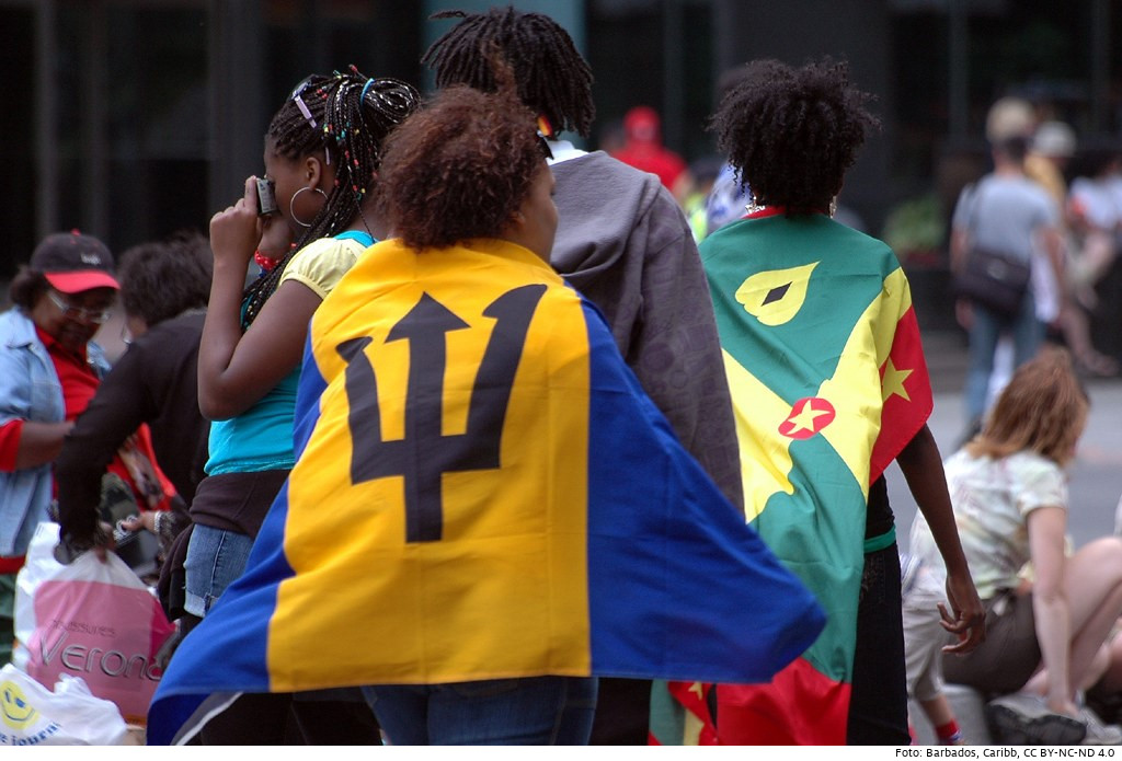 Frau im Vordergrund mit der Flagge von Barbados: Der auf gelbem Hintergrund abgebildete Dreizack des Meeresgottes Neptun soll die Verbundenheit des Inselstaats mit dem Meer symbolisieren. Foto: Barbados, Caribb, CC BY-NC-ND 4.0 